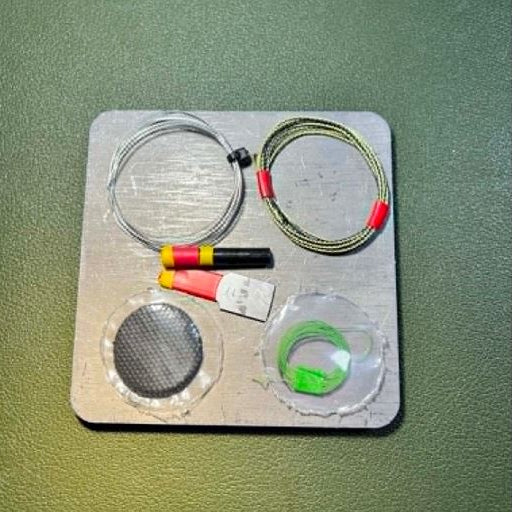 Micro‑survival hardware arranged on a metal plate, including snare wire, ferro rod components, cordage, and the char cloth disc displayed with supporting materials.