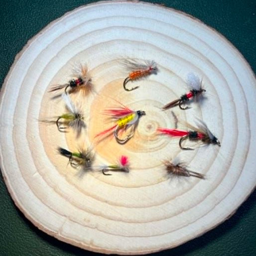 Assorted fishing flies displayed on a round wood disc, showing the patterns included in the Tiny Can of Whoop Bass Version 2.0