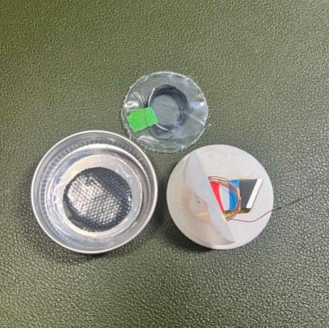 Disassembled lid components from the Tiny Can of Whoop Bass Version 2.0, including the mini knife, char cloth discs, compact suture kit, and Mylar signal mirror laid out on a green surface.