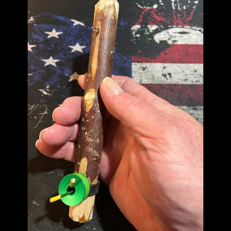 A small handmade handcaster made from a carved wooden stick with a green spool and yellow peg secured by a black band, held in a hand against a background with stars and red‑and‑white stripes.